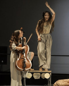 Two women in black tops and grey skirts dancing together with a long braid between them connecting each to the other. They lean away from one another.