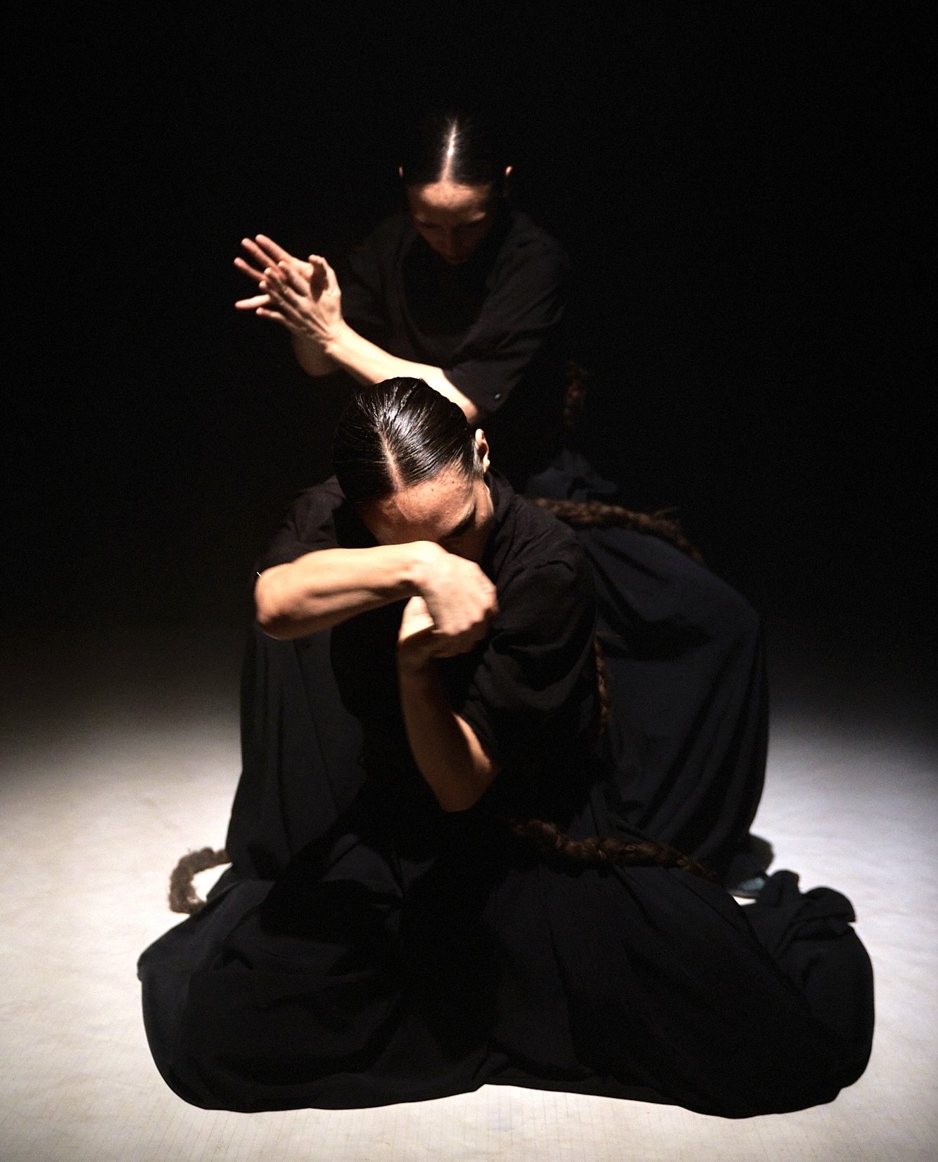 Two women in black tops and grey skirts dancing together with a long braid between them connecting each to the other. They lean away from one another.