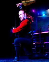 Two women in black tops and grey skirts dancing together with a long braid between them connecting each to the other. They lean away from one another.