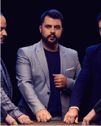 Two women in black tops and grey skirts dancing together with a long braid between them connecting each to the other. They lean away from one another.