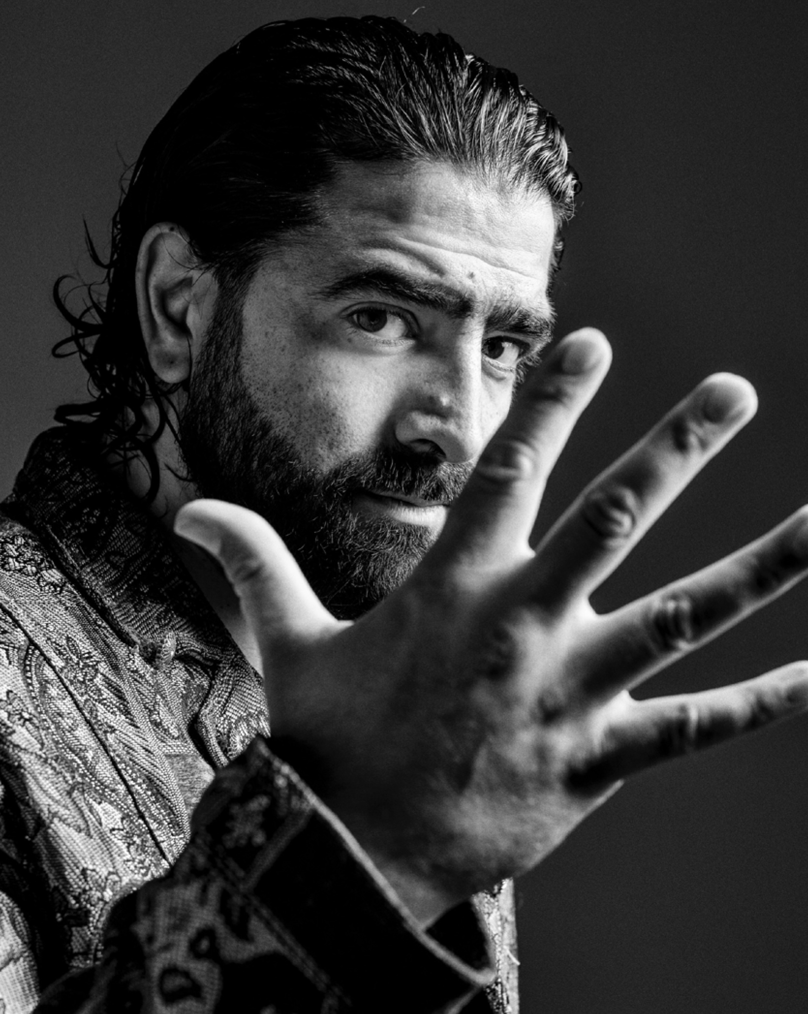 Close up black and white photo of a male flamenco dancer. He has dark hair slicked back and a beard, and the back of his hand is stretched in the photo foreground.