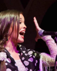 Picture of a woman singing flamenco. She has light brown hair and is gesturing with her hand outstretched by her face.